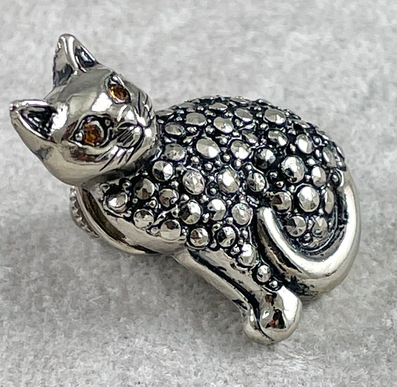 May include: A silver cat figurine with a detailed, textured surface covered in small, round stones. The cat has yellow eyes and a curled tail.