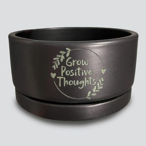 May include: A dark gray ceramic planter with a matching saucer. The planter features the text "Grow Positive Thoughts" in a light green, surrounded by a floral design. The planter is designed for indoor or outdoor use.