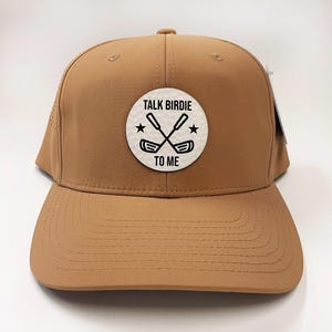 May include: A tan baseball cap with a curved brim. The front features a white circular patch with the text "TALK BIRDIE TO ME" in black, along with crossed golf clubs and stars. The cap has a structured design and a snapback closure.