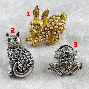 May include: Three silver and gold tone brooches. The first brooch is a rabbit with a pearl design. The second brooch is a cat with a textured design. The third brooch is a frog with a textured design.