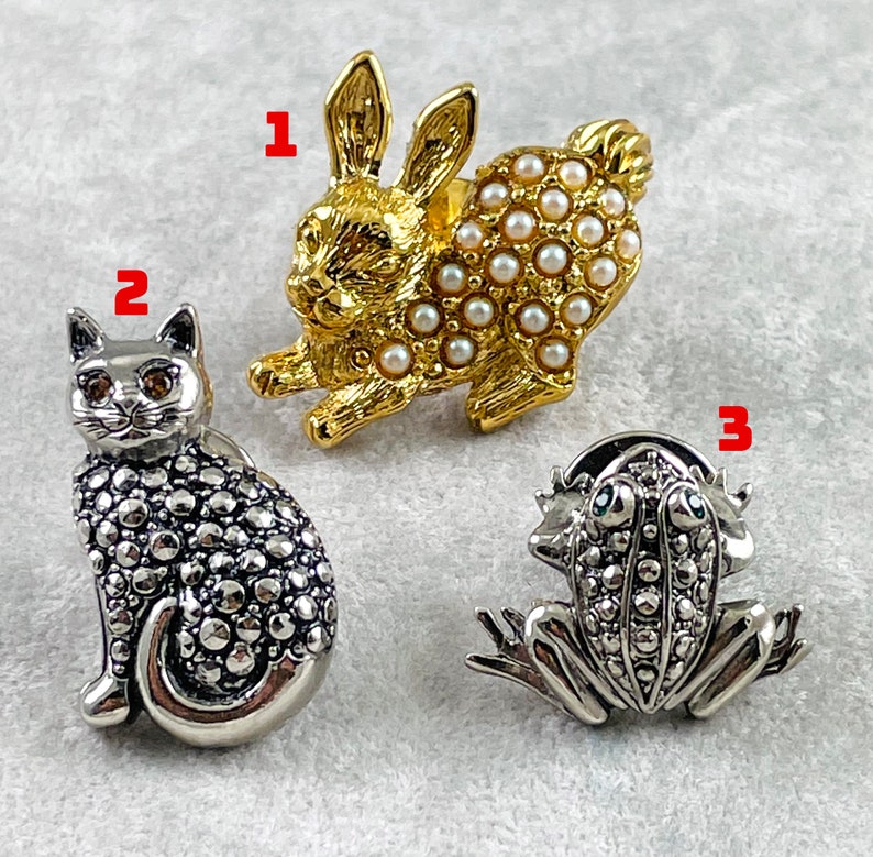 May include: Three silver and gold tone brooches. The first brooch is a rabbit with a pearl design. The second brooch is a cat with a textured design. The third brooch is a frog with a textured design.