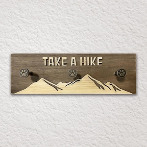 May include: Wooden wall-mounted key holder with the phrase "TAKE A HIKE" in raised letters. Features a mountain silhouette and three paw print knobs. The wood has a natural grain and a dark brown stain.