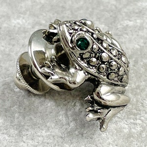 May include: A silver frog pin with a green gemstone eye. The frog is detailed with a textured surface and has a pin back.