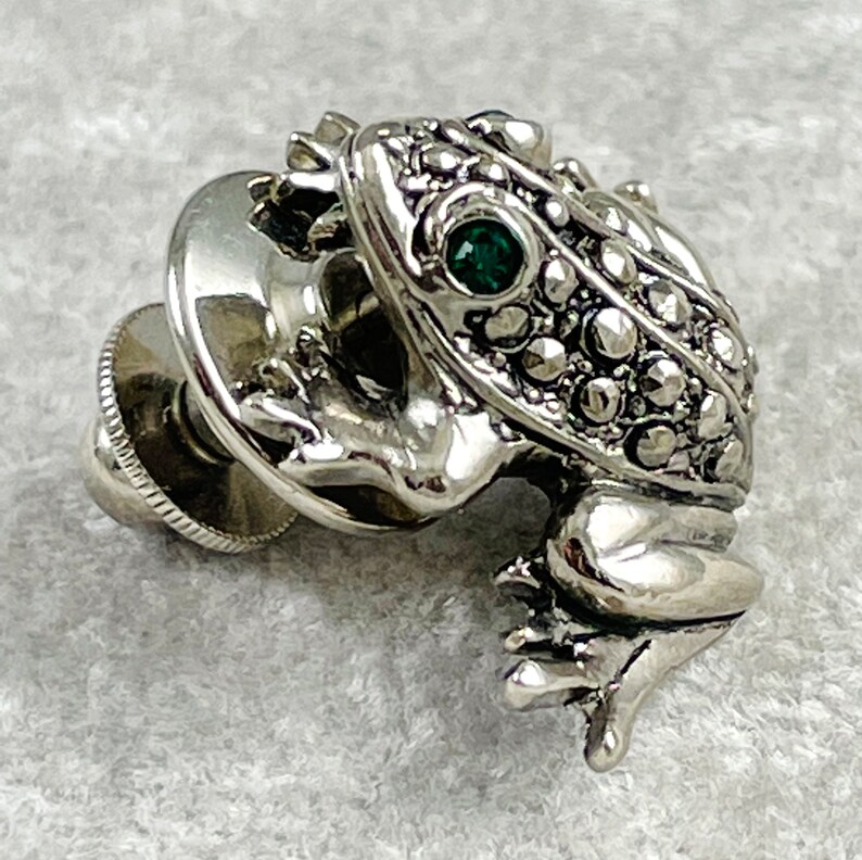 May include: A silver frog pin with a green gemstone eye. The frog is detailed with a textured surface and has a pin back.