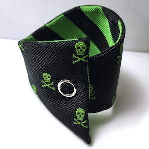 May include: Black and green fabric cufflinks with a skull and crossbones pattern. The cufflinks have a silver metal button.