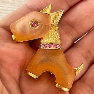 May include: A vintage dog-shaped brooch in peach and gold tones. The dog's head has a pink gemstone, and the collar is decorated with pink gems. The brooch has gold accents on the ears, tail, and feet.