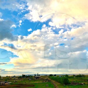 May include: A wide, open field with a blue sky and fluffy white clouds. The sky is a light blue with a few darker blue patches. The clouds are white and fluffy, and they are scattered across the sky. The field is green and grassy, and there are a few trees in the distance.