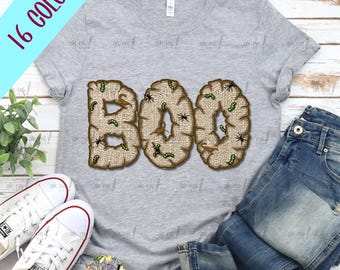 May include: A grey t-shirt featuring the word "BOO" in a burlap design, adorned with spiders and worms. The shirt is styled with blue jeans and white trainers. The text "16 COLORS" is visible in the upper left corner.