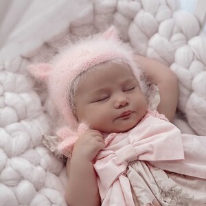 Puede incluir: Una niña bebé con un gorro rosa peludo y un vestido rosa floral con un gran lazo. La bebé está durmiendo en una manta de punto blanca.