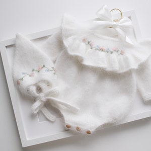 Könnte beinhalten: Weißes Baby-Set aus flauschigem Material, bestehend aus einem Strampler und einer Mütze. Der Strampler hat einen Rüschenkragen und ist mit rosa und blauen Blumenstickereien verziert. Die Mütze hat ebenfalls Stickereien und eine Schleife.