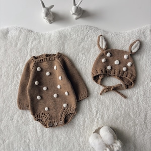 Op de afbeelding: Bruin gebreide babyromper en bijpassende muts met witte stippen. De romper heeft lange mouwen en houten knopen. De muts heeft oren en een strik. Witte konijnenbeeldjes op de achtergrond.