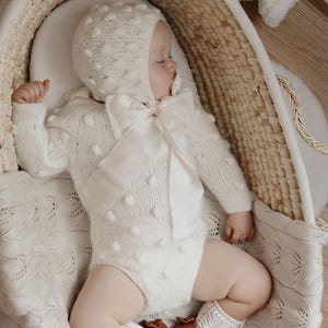 May include: A sleeping baby wearing a cream-colored knit outfit with a bonnet and a large bow. The outfit has a textured, dotted pattern. The baby is in a woven basket, wearing brown leather shoes and white lace socks. A cream-colored blanket is underneath.