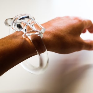 Op de afbeelding: Transparante glazen armband met een knoopontwerp. De armband wordt om de pols van een persoon gedragen. De armband is een uniek sieraad.