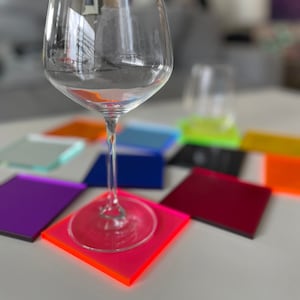 Puede incluir: Una copa de vino transparente se encuentra sobre un posavasos cuadrado rosa neón vibrante. Varios posavasos cuadrados de colores en tonos morado, azul, naranja y verde están dispuestos sobre una superficie blanca, creando una exhibición moderna y colorida.