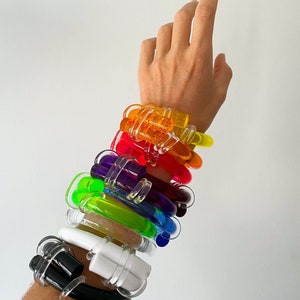 Op de afbeelding: Een stapel kleurrijke plastic armbanden in regenboogkleuren, waaronder rood, oranje, geel, groen, blauw, paars en wit. De armbanden zijn gestapeld op de pols van een persoon.