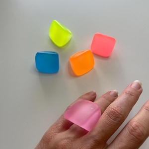 May include: A hand wearing a large, translucent pink ring. Scattered around are four other square rings in neon colors: blue, orange, pink, and yellow. The rings have a smooth, rounded shape.
