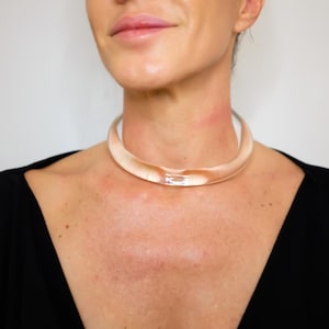 May include: A clear, light peach-colored choker necklace. The necklace is a thick, rounded tube shape with a slight curve. The model is wearing a black top. The background is a plain white wall.