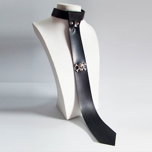 Könnte beinhalten: Eine schwarze Lederkrawatte mit einem silbernen Totenkopf- und Knochen-Akzent. Die Krawatte wird auf einer weißen Puppe präsentiert.