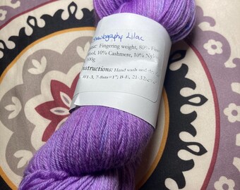 Può includere: Un gomitolo di filato viola con un'etichetta bianca che recita "hawography Lilac Fingering weight, 80% Fine Wool, 10% Cashmere, 10% Nylon 100g Instructions: Hand wash and dry flat #1-3, 7-8sts=1" B-E, 21-32"