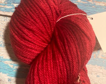 Puede incluir: Una madeja de hilo rojo vibrante, bien enrollada y lista para tejer. El hilo es de un color rico y sólido, con una textura suave. Una etiqueta blanca está adjunta. El fondo es una superficie de madera pintada y desgastada.