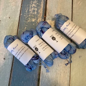 May include: Four skeins of blue and purple speckled yarn with white labels. The labels read "Kitty Bennet" and include yarn details. The yarn is arranged on a weathered, blue-painted wooden surface.