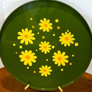 May include: A round, green metal tray with a gold rim and gold stand. The tray is decorated with yellow daisies and orange dots. The tray is a vintage piece, perfect for serving or display.