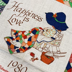May include: A white fabric calendar for 1980 featuring a brown embroidered phrase Happiness is Love. The design shows a figure in a blue hat sitting in a rocking chair with a colorful patchwork quilt, a white cat, and a brown woven basket.
