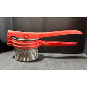 May include: A vintage red potato ricer with a perforated stainless steel base and long handles. The kitchen utensil shows signs of wear, with chipped paint and a well-used appearance. The ricer is designed for mashing potatoes and other soft foods.