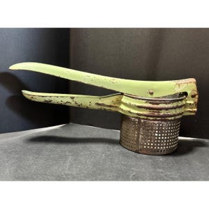 May include: Vintage potato ricer with a light green handle and a perforated metal base. The handle shows signs of wear, with chipped paint. The ricer is designed to press cooked potatoes, creating a smooth texture. A classic kitchen tool.