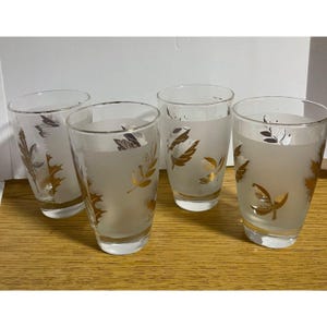May include: A set of four clear drinking glasses with a frosted finish and metallic gold leaf patterns. Each glass features a tapered cylindrical shape with a smooth rim and a clear base, decorated with various leaf designs in gold.