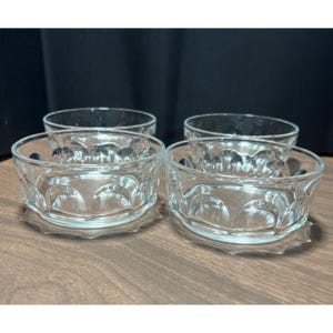 May include: Four clear glass bowls with a decorative pattern around the sides and a scalloped base. The bowls are arranged on a wooden surface, with a dark background.