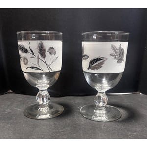 May include: Two clear wine glasses with frosted white bands featuring floral and leaf designs in black and gray. The glasses have a silver base and stem with a decorative bulb detail. The background is dark.