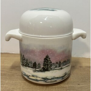 May include: A white ceramic container with a lid and two handles. The container is decorated with a winter landscape scene featuring trees, snow, and a pink and purple sky. The lid is rounded and fits snugly on top.