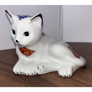 May include: A white ceramic cat figurine in a resting pose. The cat has black eyes, ears, and paw pads. It is decorated with floral patterns in red, blue, and gold. A decorative item for cat lovers.