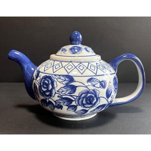May include: A white and blue ceramic teapot with a floral design. The teapot features a blue handle, spout, and lid. The body of the teapot is white with blue floral patterns and geometric shapes. The lid has a small blue knob.