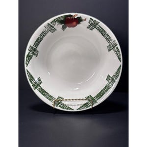 May include: A round white ceramic bowl featuring a green cross-hatched fence pattern around the inner rim and a single red apple illustration with green leaves positioned at the top center of the interior surface of the bowl.