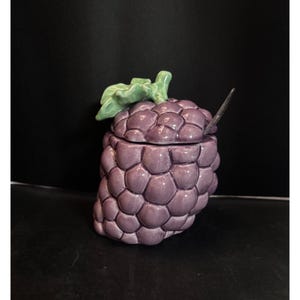 May include: A ceramic jar shaped like a cluster of purple grapes, with a matching lid and a small spoon. The lid is topped with a green leaf. The jar is a decorative kitchen item.