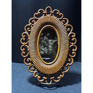 May include: A decorative oval mirror with a brown, ornate frame. The frame has a textured, ribbed design around the mirror's edge and decorative scrollwork. The mirror is supported by a silver metal stand.