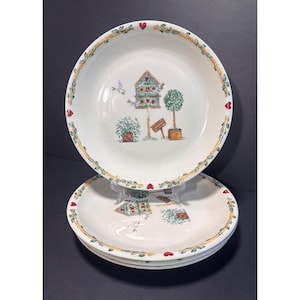 May include: A set of three white ceramic plates featuring a central illustration of a blue birdhouse, a green topiary, and small birds. The rim is decorated with a yellow vine pattern interspersed with small red hearts around the edge.