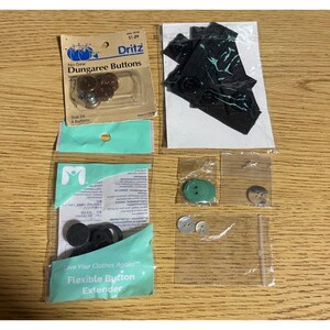 May include: A collection of sewing notions including a package of Dritz No-Sew Dungaree Buttons, a black fabric button extender, a package of Flexible Button Extender, a small green button, a translucent button, and two small white buttons.