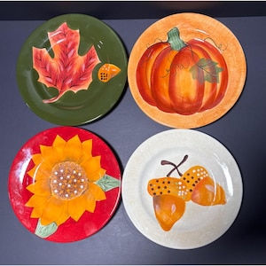 May include: Four decorative ceramic plates with fall-themed designs. One plate features a red and orange maple leaf on a green background. Another has a large orange pumpkin. A sunflower is on a red plate, and acorns are on a cream-colored plate.