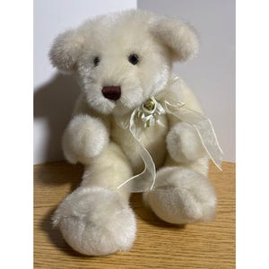 May include: A plush teddy bear with cream-colored fur, dark eyes, and a dark brown nose. The bear is sitting in an upright position and features a translucent, off-white organza ribbon tied in a bow around its neck, accented with a small green bead.