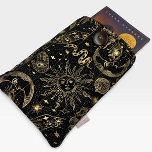 May include: A black fabric book sleeve featuring a gold celestial print of suns, moons, stars, snakes, and hamsa hands. The quilted pouch has a dark brown button closure and contains a book with the text Frank Herbert Dune Part Two visible.