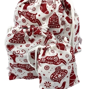 May include: Three off-white drawstring bags of varying sizes featuring a repeating red print of reindeer, bells, hearts, stockings, birds, and snowflakes. Each bag is made of fabric and includes a white cord closure at the top for securing contents.