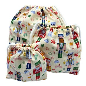 Puede incluir: Tres bolsas con cordón con un fondo crema y un patrón repetido de coloridos cascanueces, árboles de Navidad y copos de nieve. Las bolsas tienen cordones blancos y varían en tamaño.