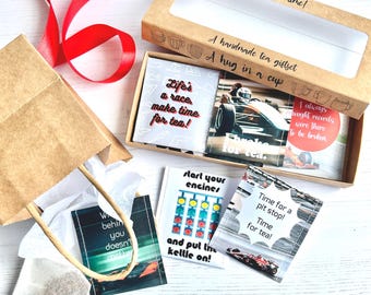 Può includere: Una borsa regalo di carta marrone con una fodera in carta velina bianca e una bustina di tè all'interno. La borsa è accanto a una scatola che contiene un set regalo di tè a tema corse. La scatola ha una finestra trasparente e il testo "It's tea time! A handmade tea giftset A hug in a cup" è stampato sul davanti. Le bustine di tè hanno disegni a tema corse e motti come "Life's a race make time for tea!" e "Time for a pit stop! Time for tea!"