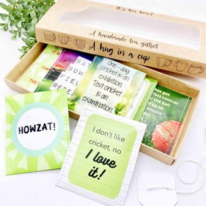 May include: A handmade tea gift set in a brown box with a clear window. The set contains tea bags and cards with cricket-themed quotes. The box is labelled "A hug in a cup". The cards include phrases like "Howzat!" and "I love it!"