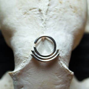 May include: A silver septum ring with three stacked, concentric circles. The ring is shown on a white, textured surface.