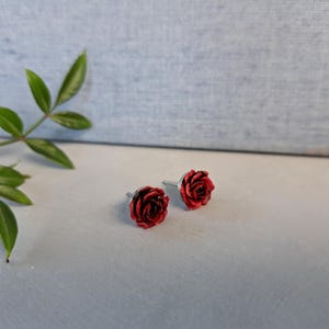 Może przedstawiać: Para kolczyków typu sztyfty z detalami w kształcie ciemnoczerwonych róż. Każda róża jest przymocowana do srebrnego metalowego sztyftu, prezentując skomplikowane warstwy płatków i realistyczny kwiatowy wzór tych małych akcesoriów.
