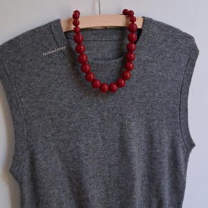 Pu&ograve; includere: Un gilet in maglia grigio senza maniche con scollo rotondo, caratterizzato da una collana di grandi perle rosse rotonde e lucide drappeggiata sul colletto. Il testo karavakishop &egrave; stampato in piccole lettere bianche sul lato superiore sinistro.